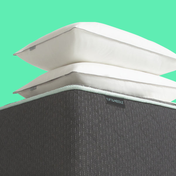 A close-up of a Tuft and Needle Original mattress corner with two Tuft and Needle stacked white pillows, showing a dark gray textured fabric cover and a small visible Tuft and Needle label, against a bright mint green background.