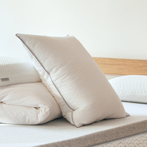 Beige Tuft and Needle pillows and folded Tuft and Needle Duvet bedding sitting on top of a mattress protector that is on top of Tuft and Needle mattress on a bed frame with a wooden headboard.