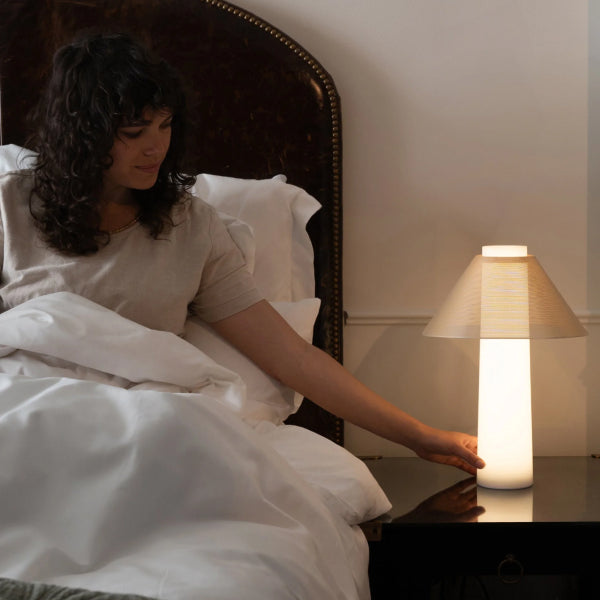 A woman in bed reaches toward a glowing cylindrical table lamp on a bedside table. The Tuft and Needle Loftie Lamp has a warm beige shade and emits a soft white light, set in a bedroom with white bedding and a dark upholstered headboard.