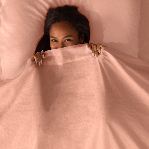Woman laying on a tuft and needle mattress with her had on a pillow in and her head half covered underneath pink tuft and needle bedding