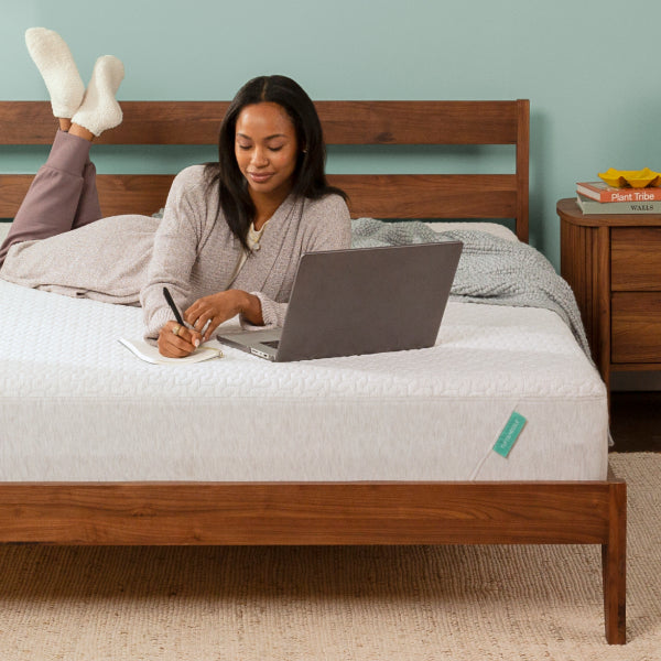 Woman laying on top of Tuft and Needle mattress in a brown bedframe writing on a pad to the right of her laptop.