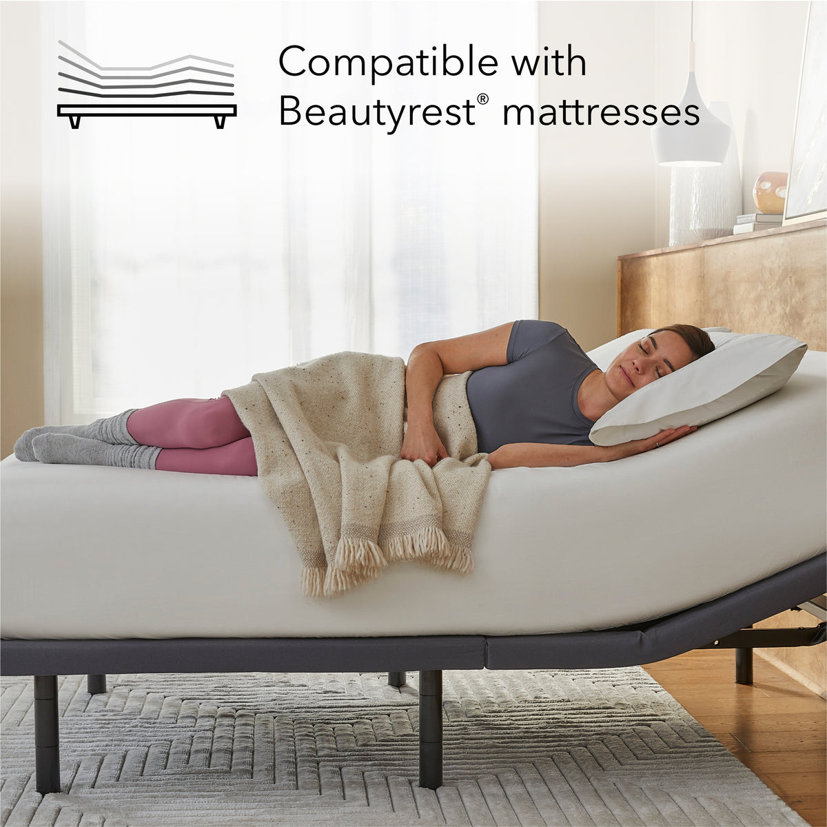 Woman lying on a Beautyrest mattress on top of a Pearl adjustable base with a pillow, surrounded by a blanket, in a bedroom setting.