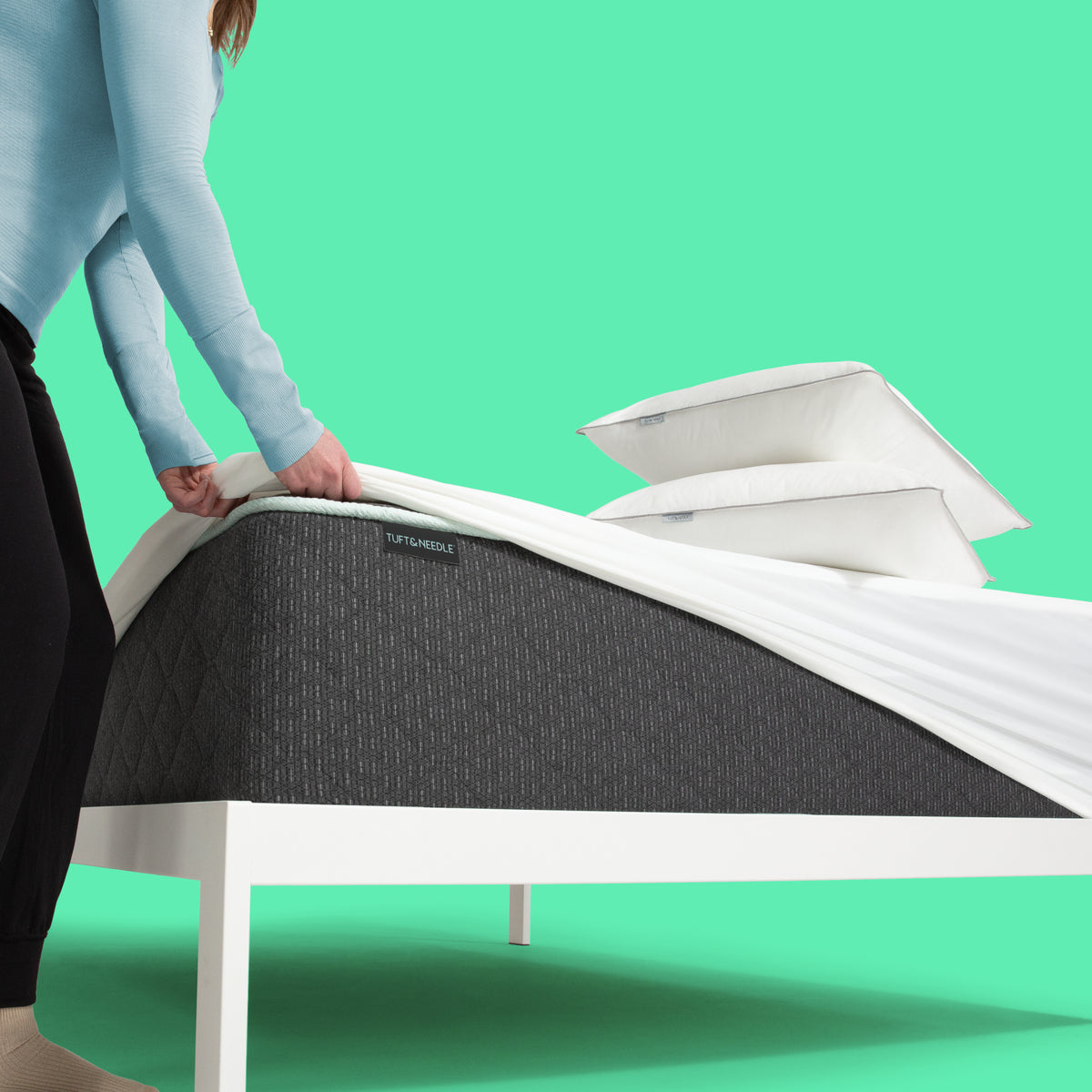 A woman pulling a mattress protector over a Tuft and Needle Mint mattress II with pillows stacked.